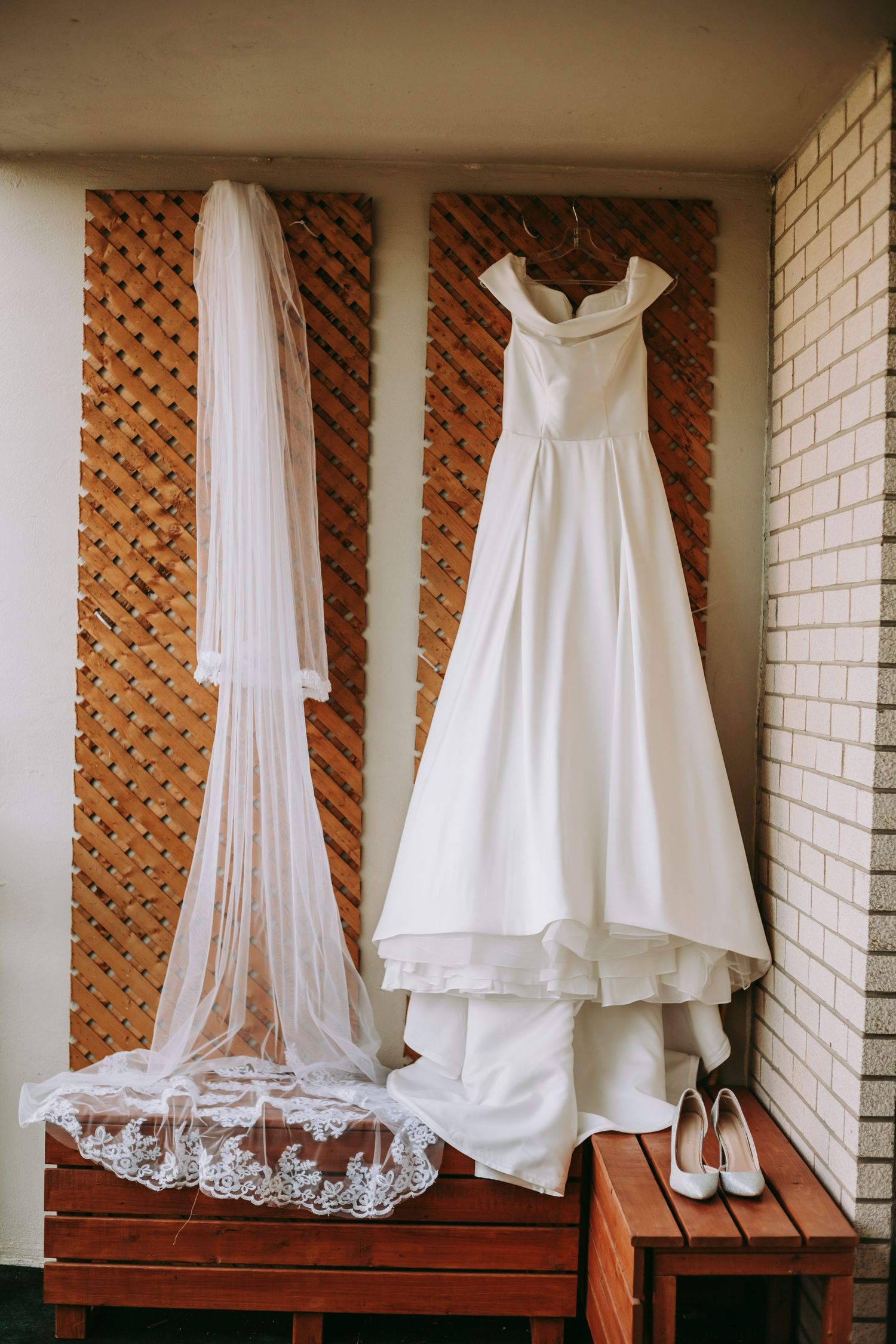 Elegant white wedding dress and veil hanging against lattice wall with bridal shoes.