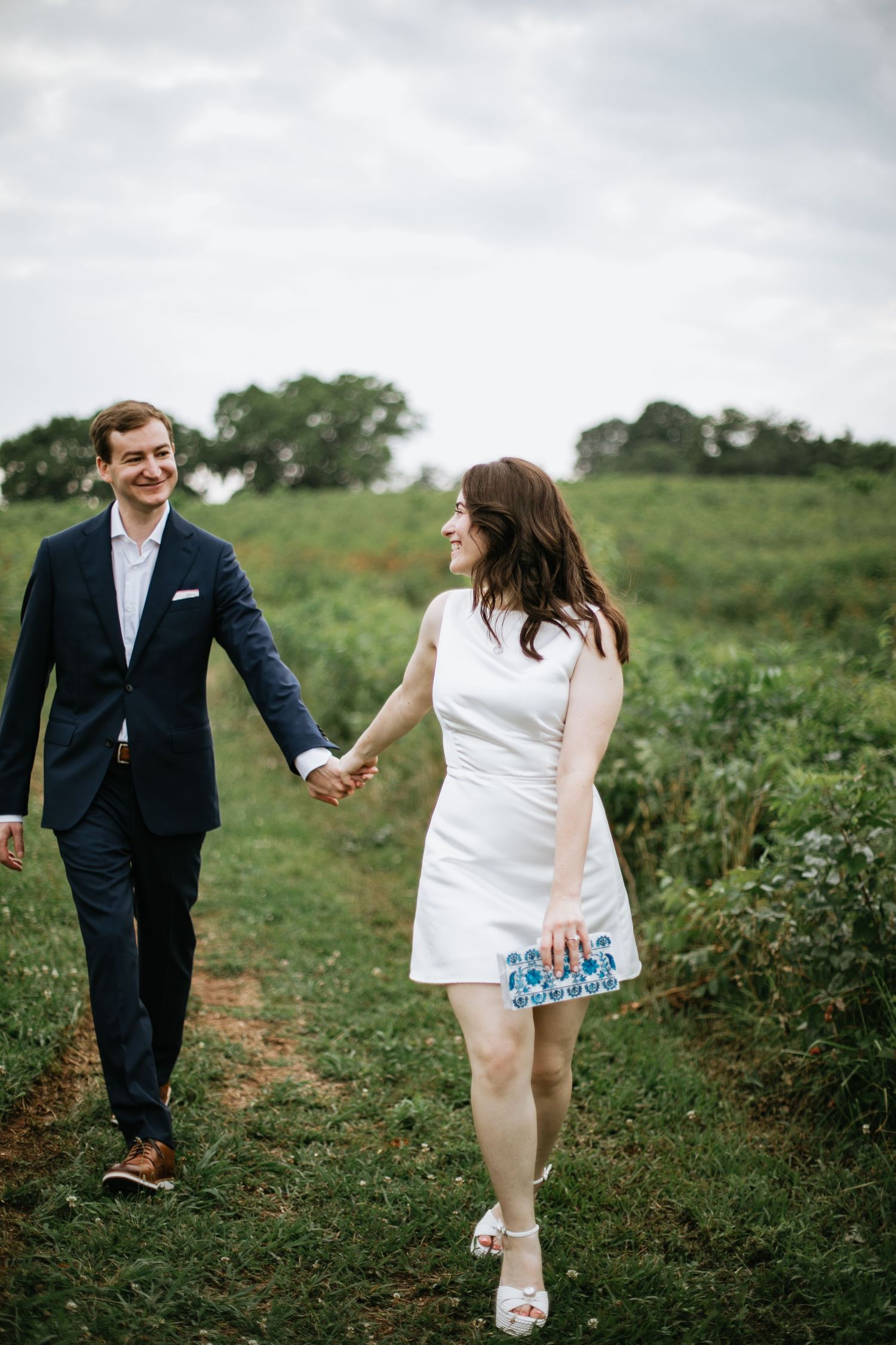 A couple in wedding attire walks through a green field on a cloudy day.