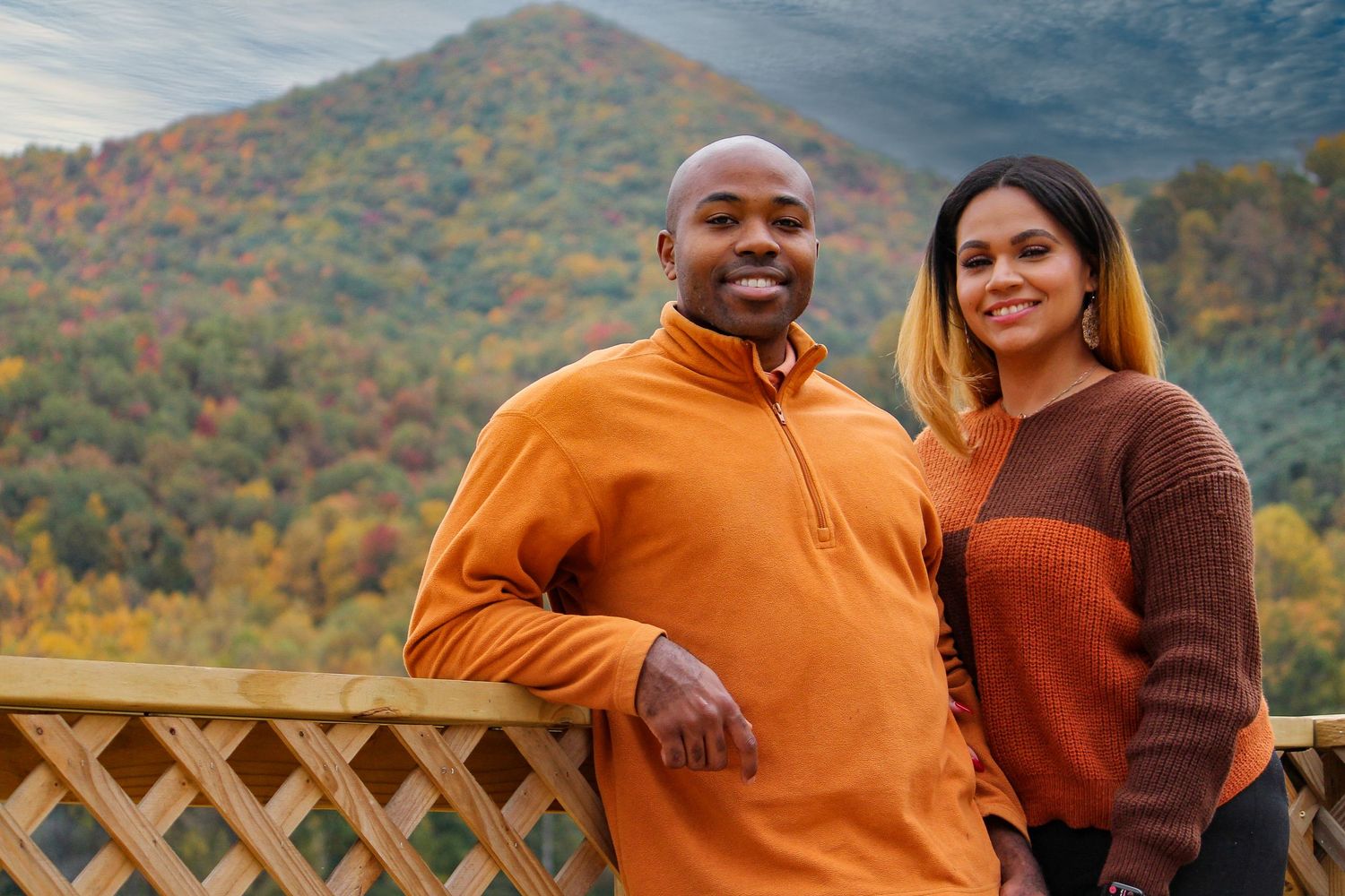 A couple stands together on a deck overlooking autumn mountains wearing orange and rust-colored sweaters.