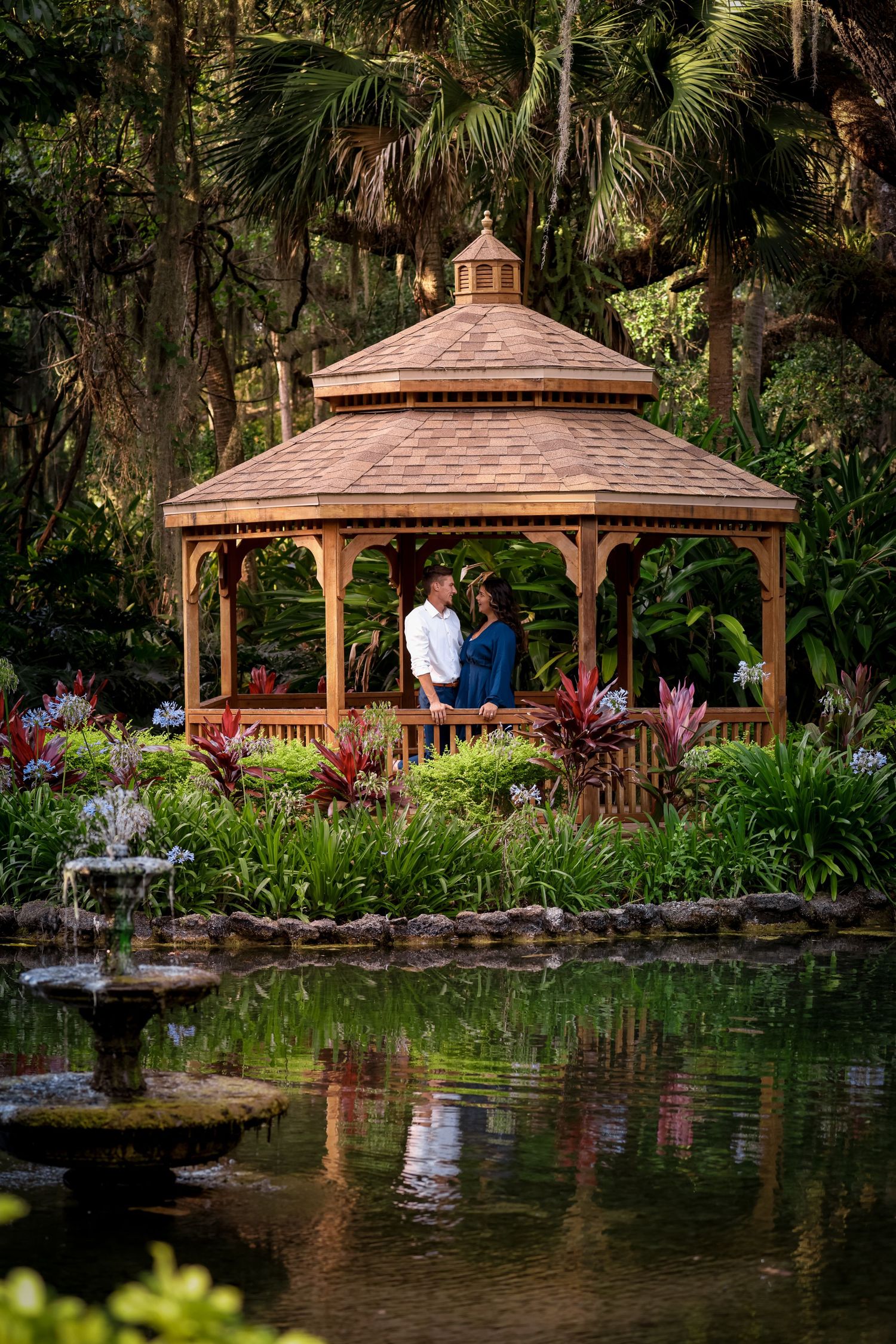 A romantic wooden gazebo overlooking a tranquil pond surrounded by tropical plants and palm trees in a lush garden.