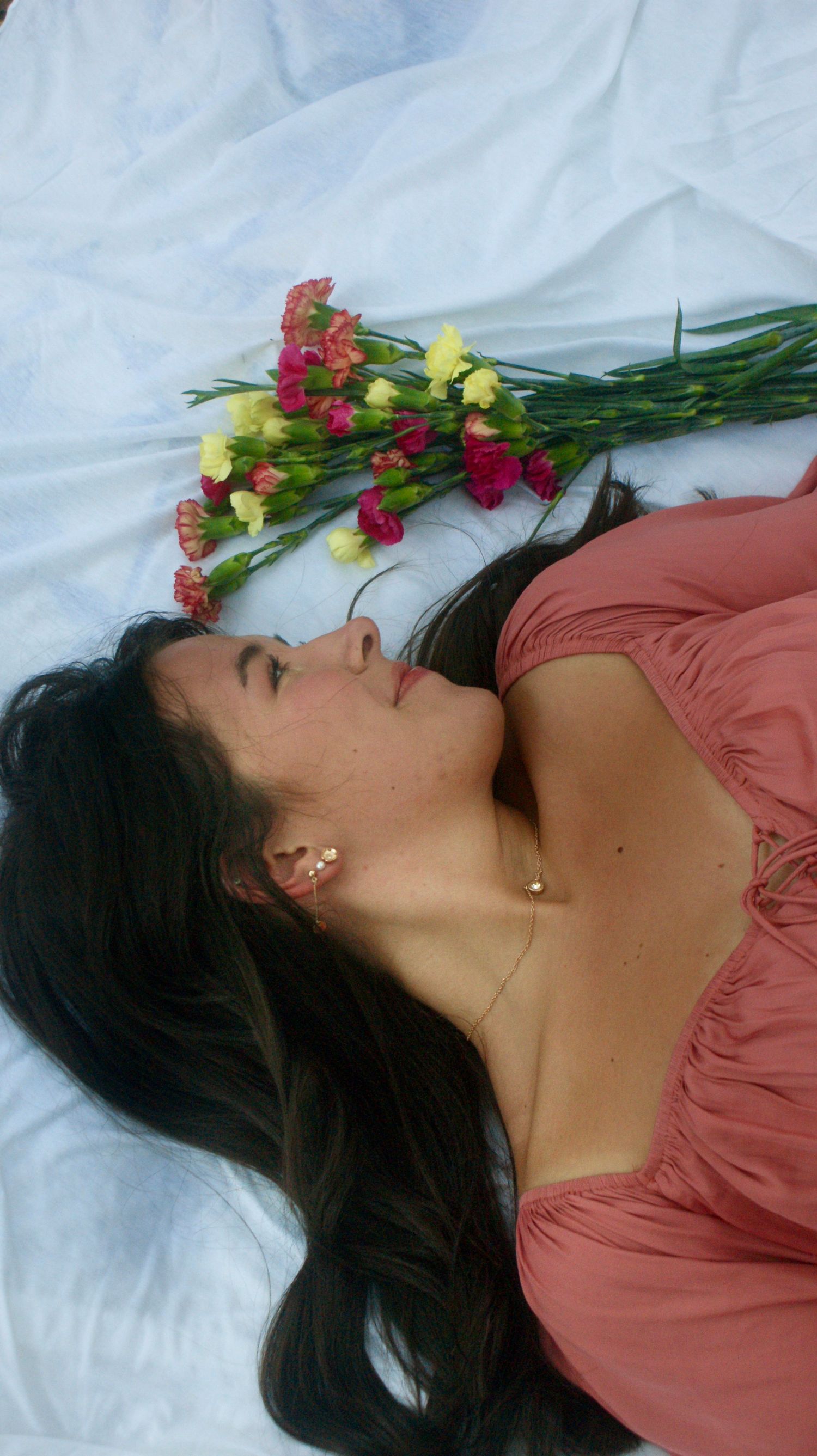 Person in coral dress lying on white fabric with colorful flowers placed near their head.