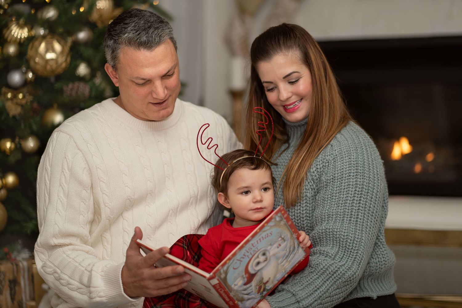 Family shares tender moment while reading book together beside fireplace and Christmas tree.