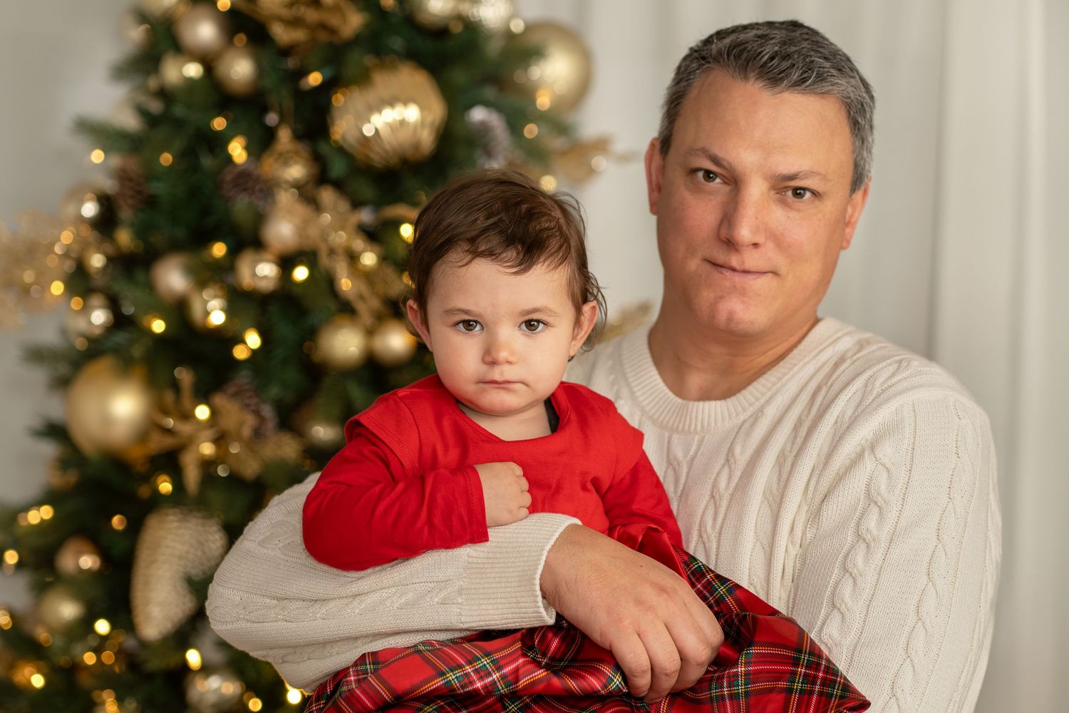 Father and child in holiday attire pose in front of decorated Christmas tree.