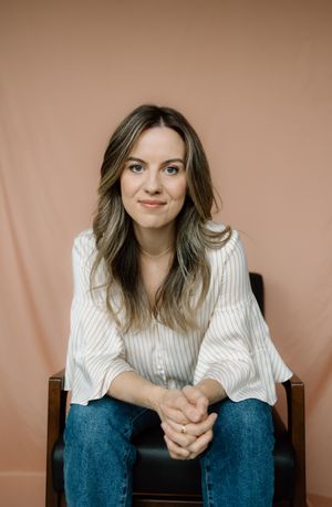 Professional studio portrait of person in white striped shirt and jeans sitting casually against peach backdrop.