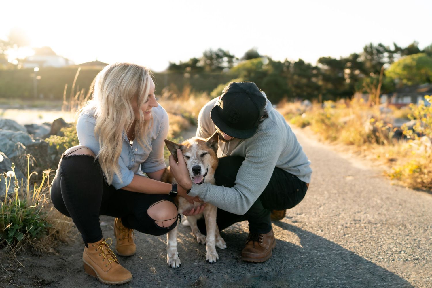 A couple petting their dog on a sunlit path during golden hour.