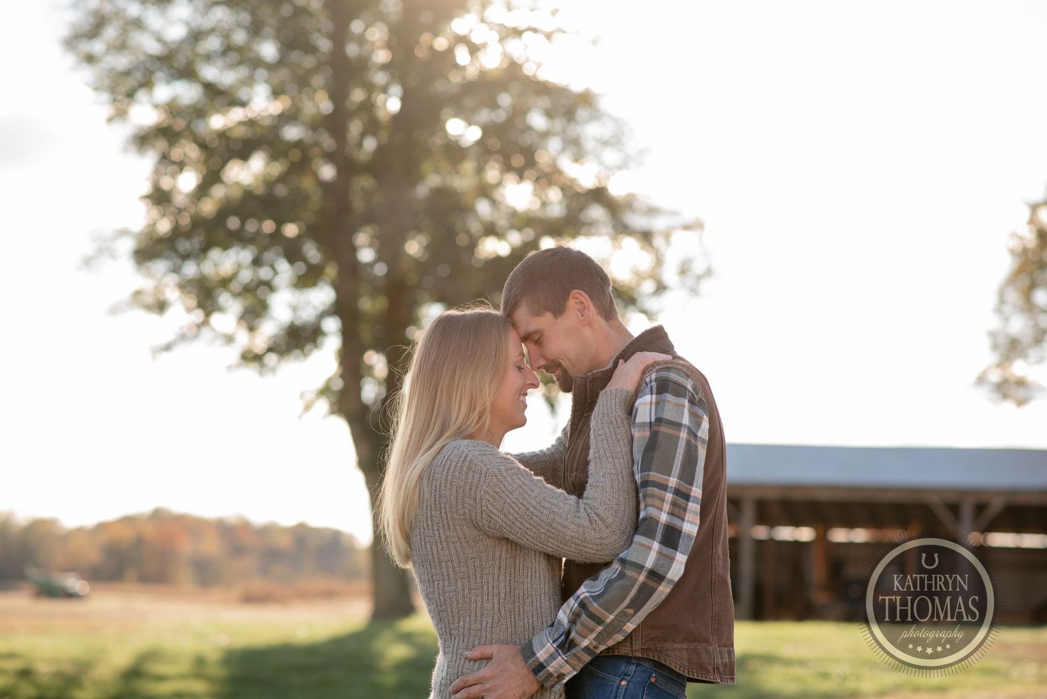 Couple embracing in golden sunset light with barn and trees in background.