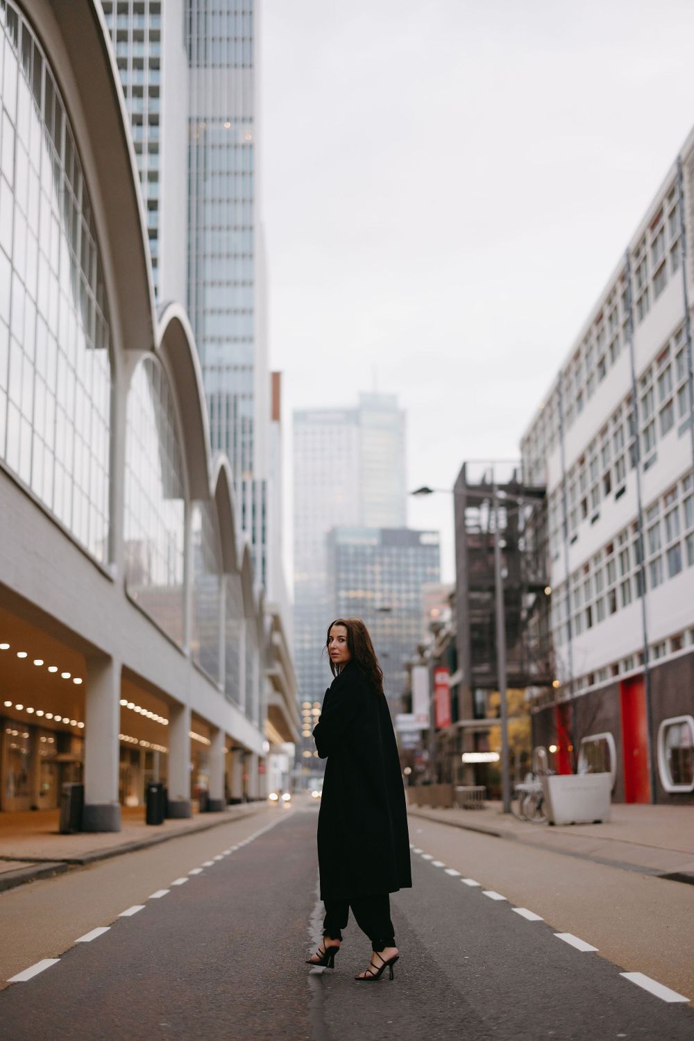 Stylish portrait of someone in a long black coat standing in an urban street with modern architecture.