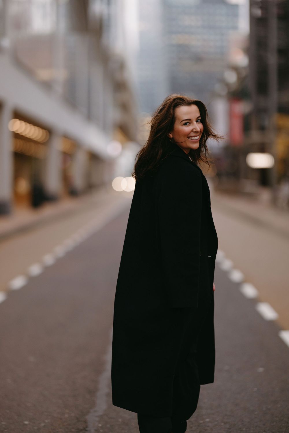 Candid street style photo capturing laughter in long black coat against urban backdrop.
