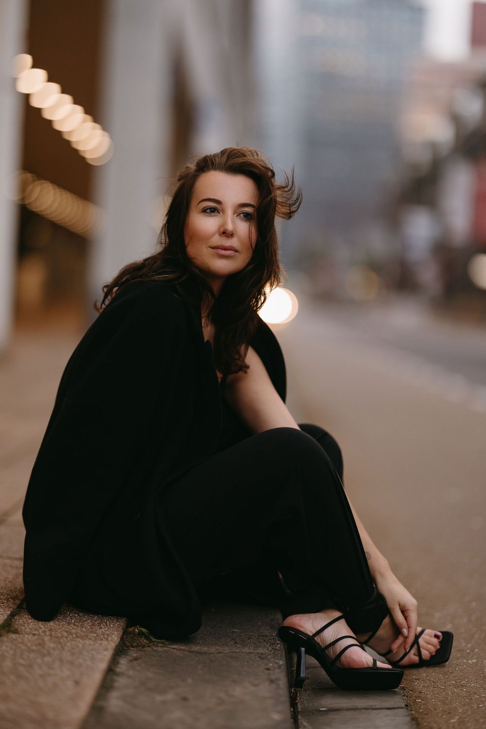 Fashion photo of someone in all black ensemble sitting on city sidewalk with bokeh lights.