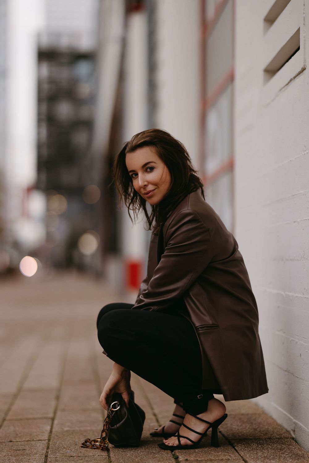 Candid fashion portrait crouching by white wall wearing brown blazer and strappy heels.