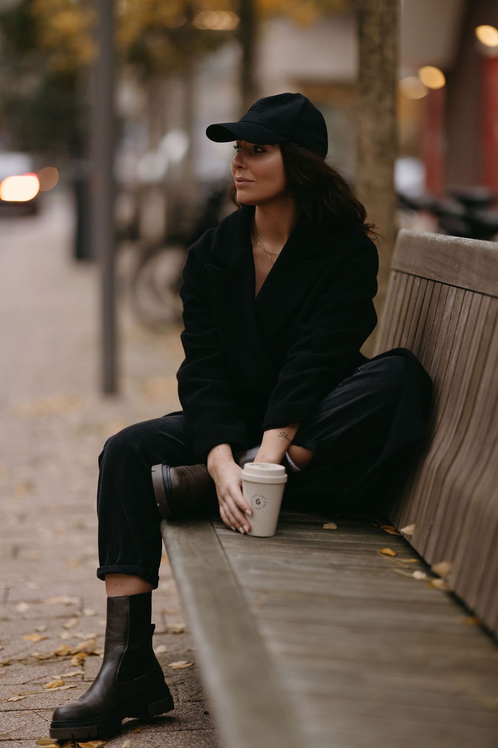 Casual autumn street style with black coat, baseball cap and combat boots while holding coffee.
