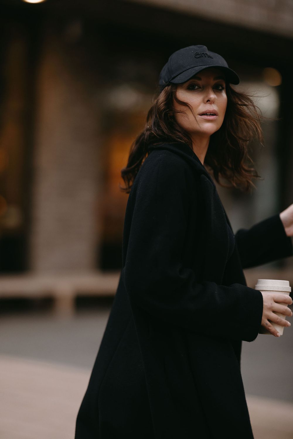 Urban street style featuring black coat and baseball cap while holding coffee on city sidewalk.