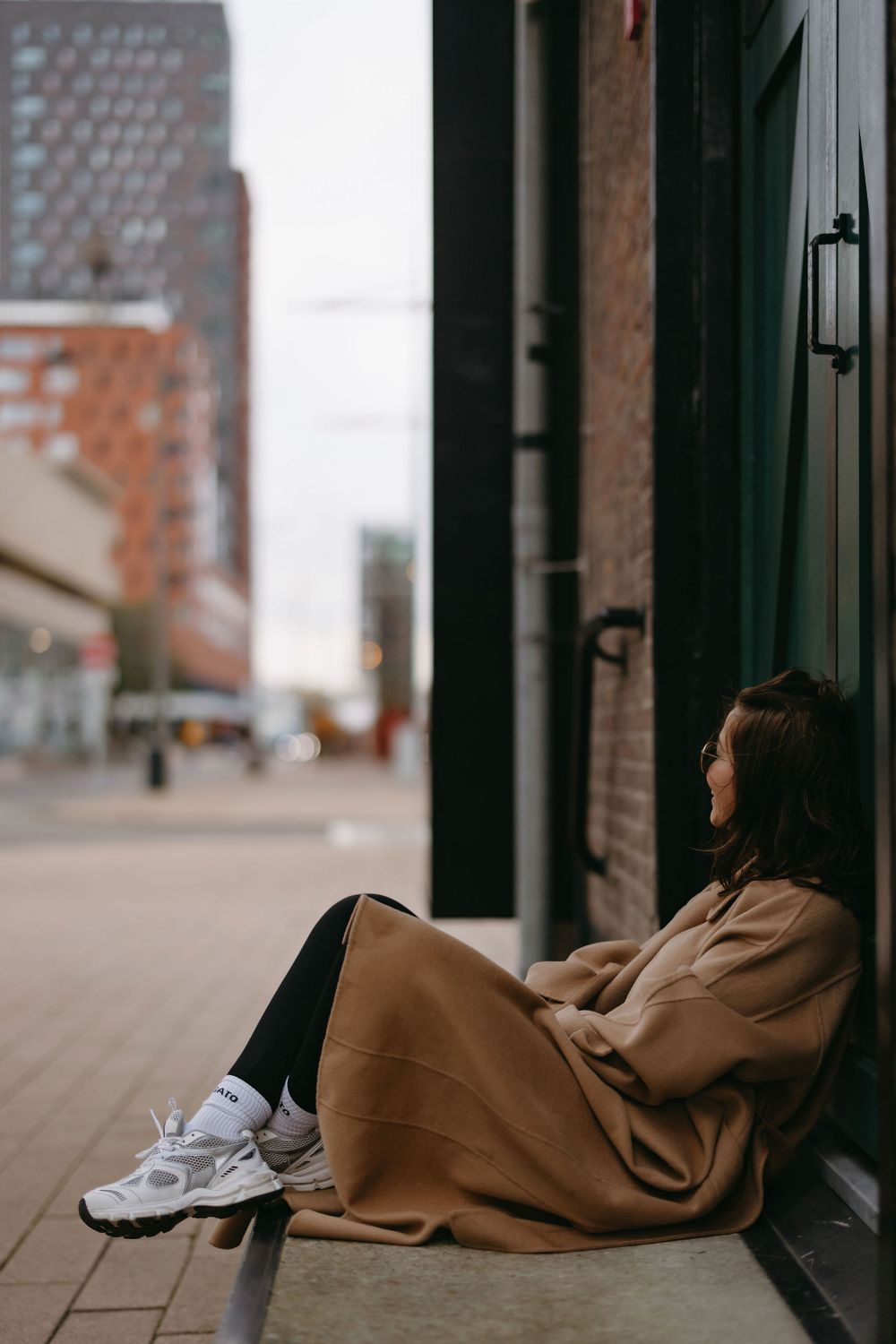 Relaxed street fashion scene with person in camel coat sitting against urban building.