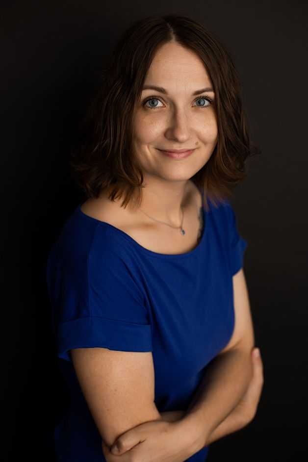 Woman in blue dress and black backdrop standing angled towards light with arms crossed.