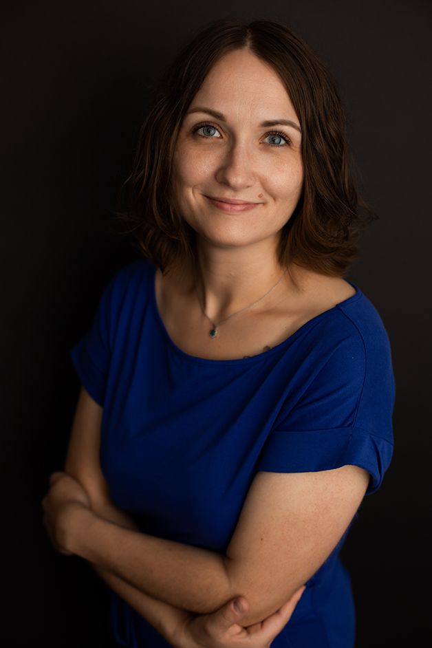 Woman in blue dress with black backdrop facing away from light with arms crossed.