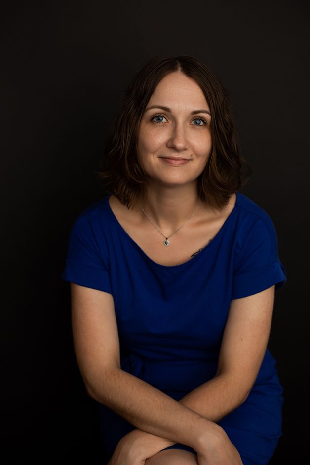 Woman sitting with arms on her knees with black backdrop looking at camera smiling.
