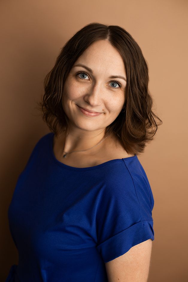 Woman on brown backdrop wearing a blue dress angled away from camera smiling.
