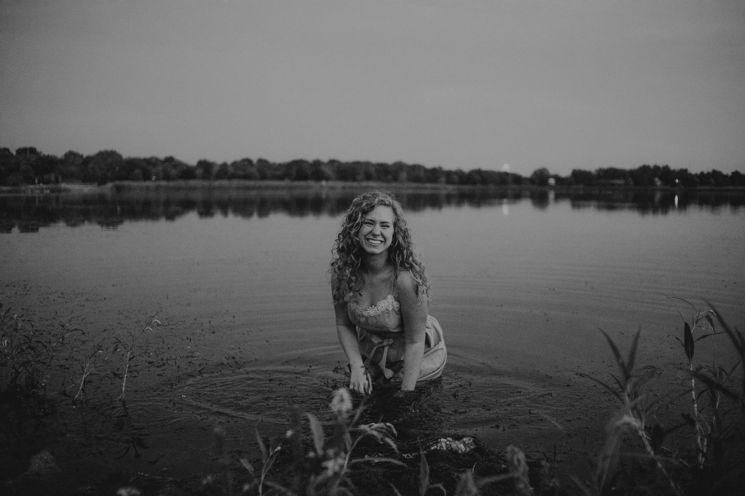 Black and white photo of woman sitting by tranquil lake at sunset.