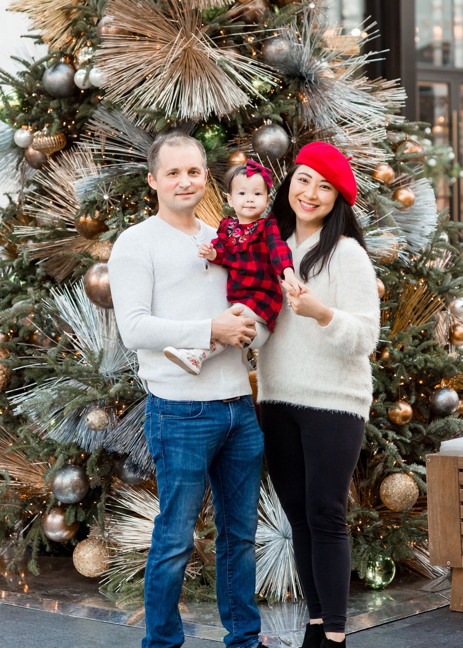 family of three taking christmas mini portraits in front of a decorated christmas tree wearing a red plaid dress