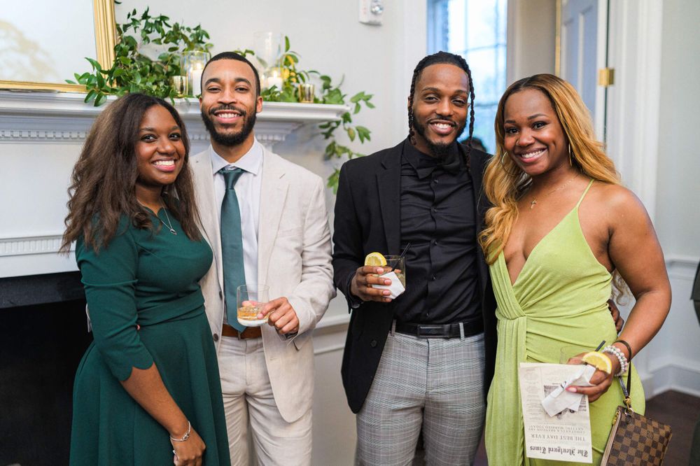 Four wedding guests pose together in formal attire against a white backdrop with greenery.