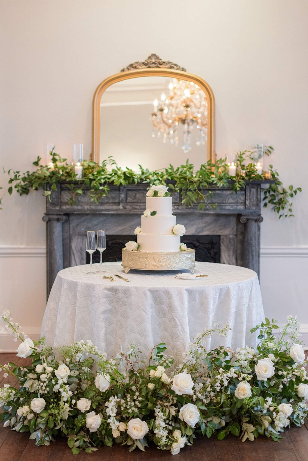 Classic white wedding cake display with cascading greenery and roses around an ornate gold mirror.