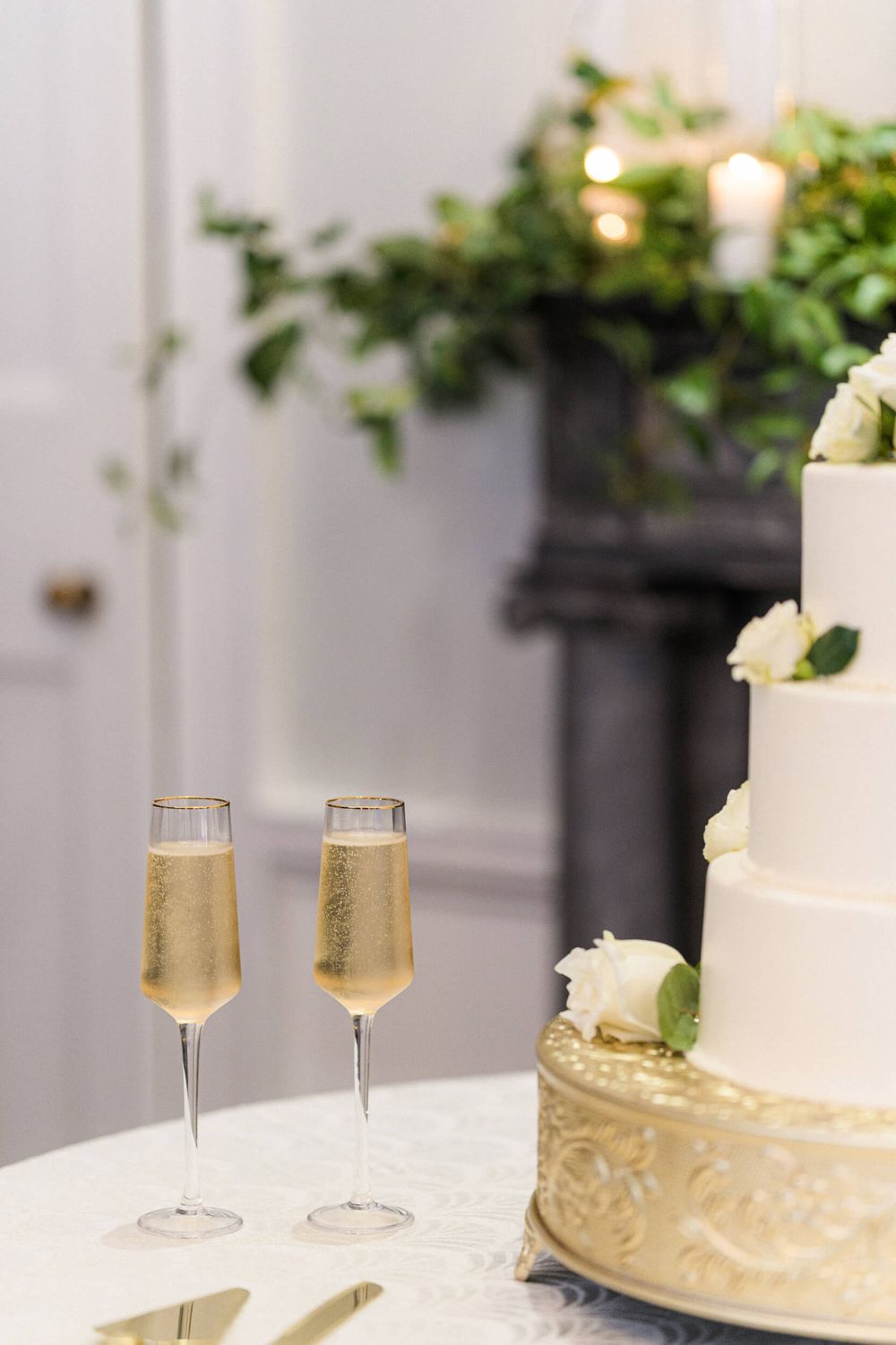 Elegant wedding cake decorated with white flowers sits beside two champagne flutes on a white table.