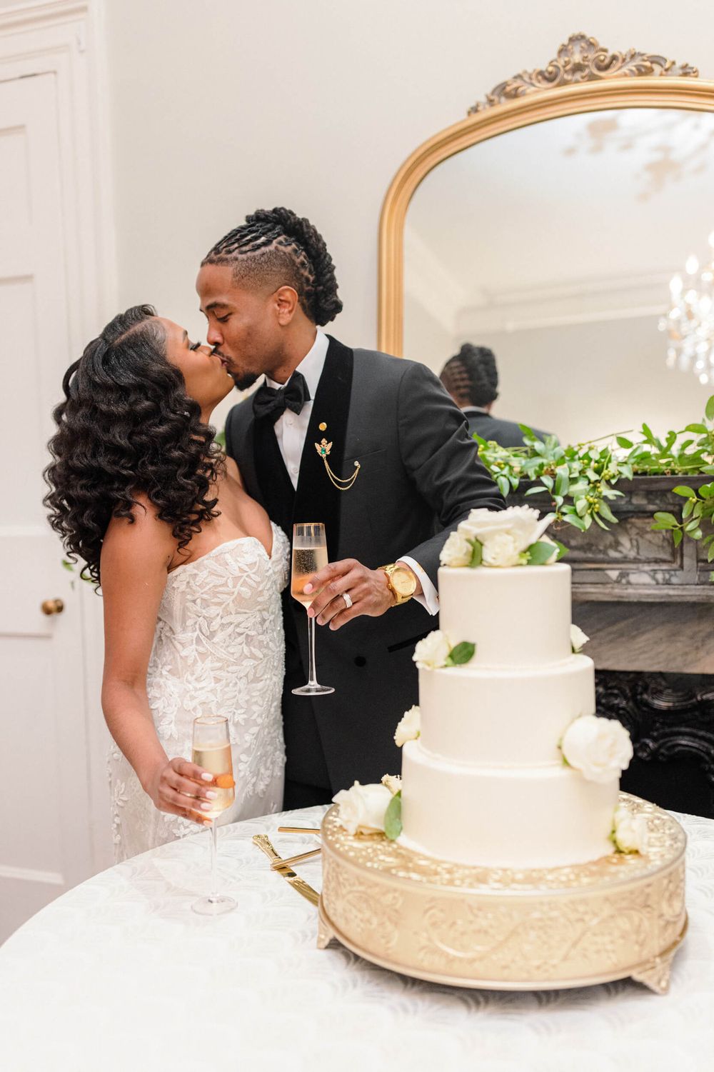 A wedding couple shares a romantic kiss by their elegant white and gold tiered wedding cake.