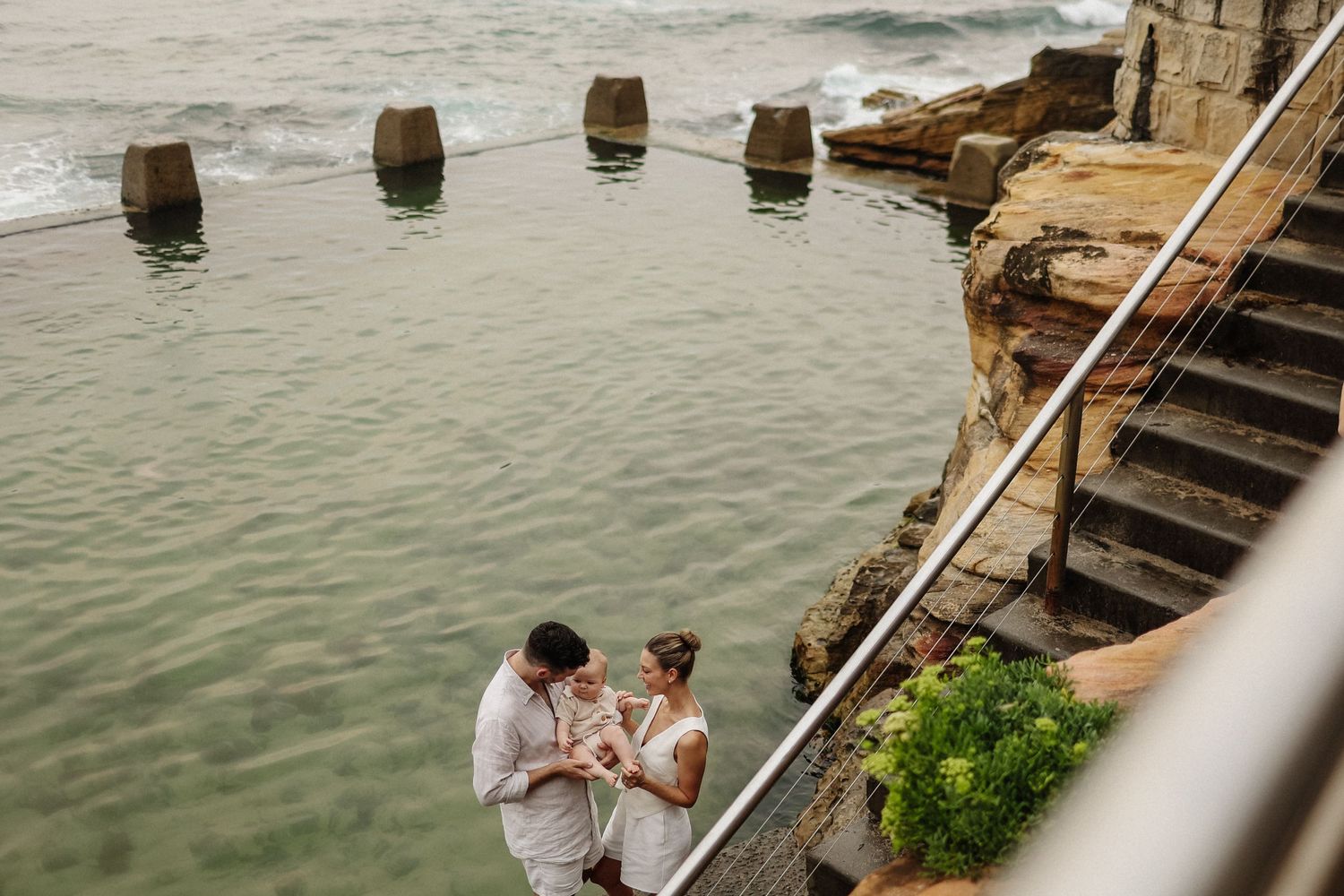 Couple sharing a tender moment beside a coastal ocean pool with stone steps behind them.