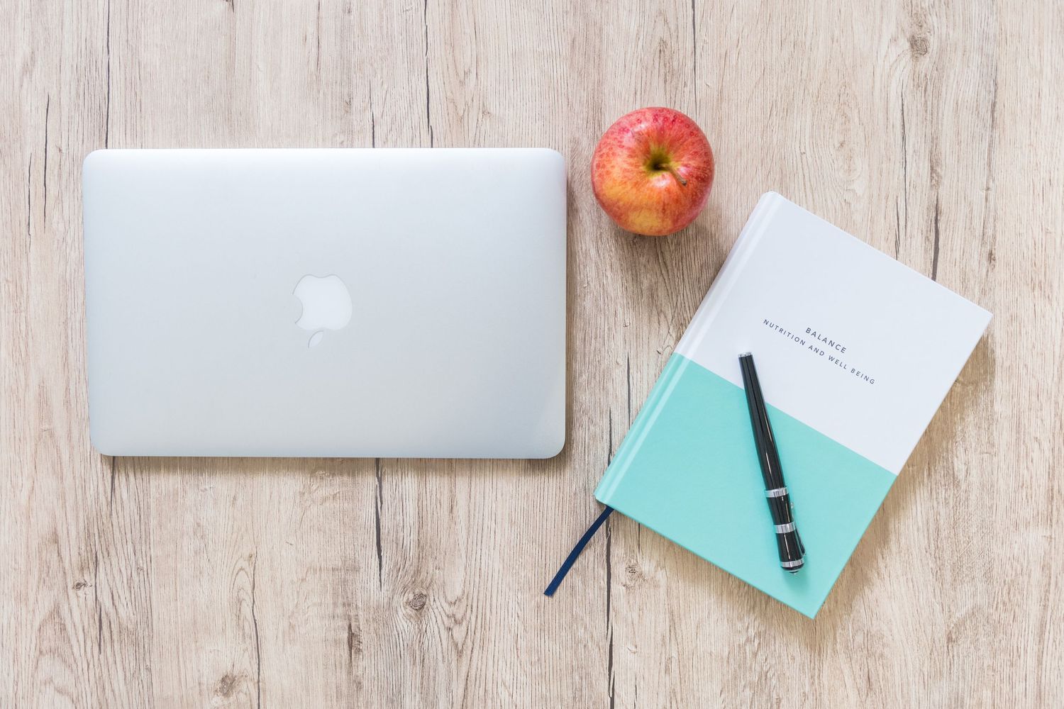 Minimalist desk setup with apple, laptop and writing materials on white wooden surface.