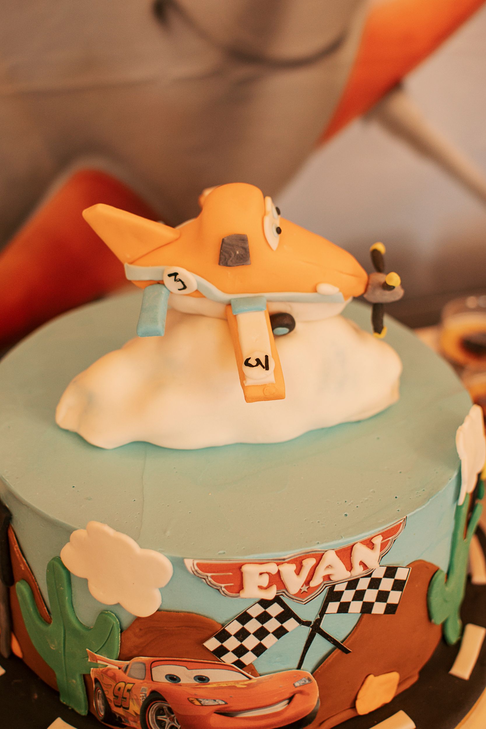 Cars themed birthday cake with orange and teal decorations featuring checkered flags and racing motifs.