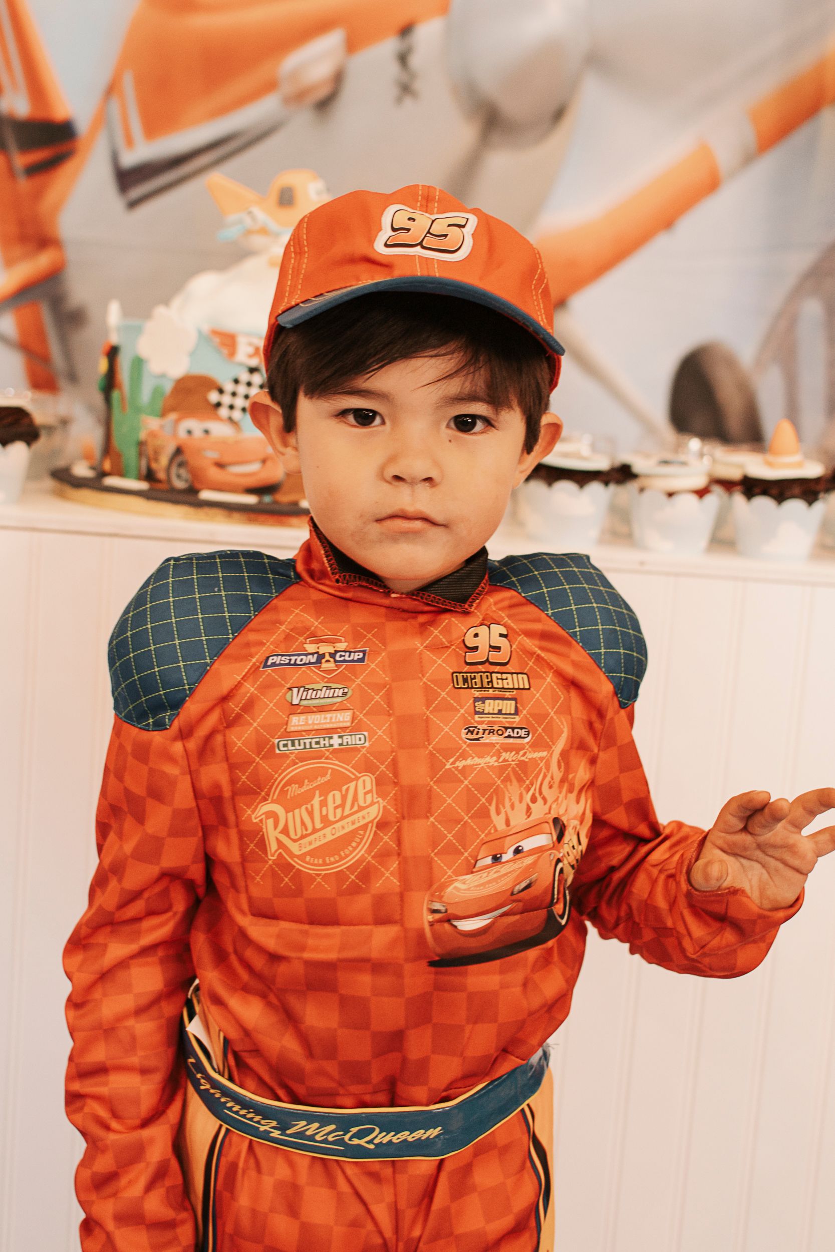 Child wearing orange racing costume with checkered pattern and baseball cap.