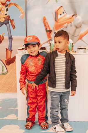 Two children stand together at a party, one wearing a red outfit and baseball cap.