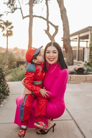 A heartwarming moment captured outdoors with bright pink and red outfits against a backdrop of palm trees and stone walls.