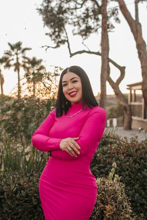 Woman in a fitted hot pink dress poses confidently outdoors at sunset with palm trees in background.