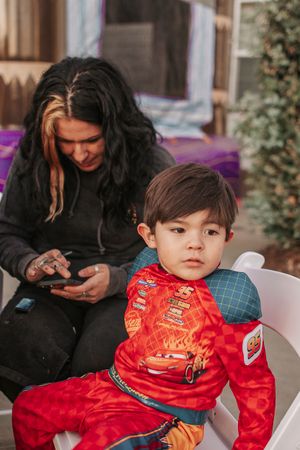 Child wearing a red Cars-themed outfit sits in a chair while caregiver looks at phone.