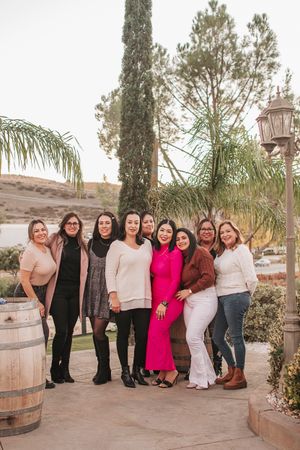Group photo of women in casual attire posing together on a patio with wine barrels and cypress trees.