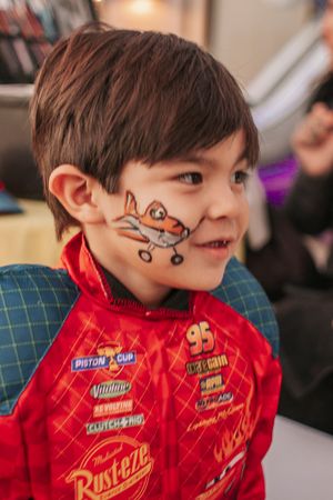 Young child with face paint wearing a red Cars themed shirt smiles brightly.