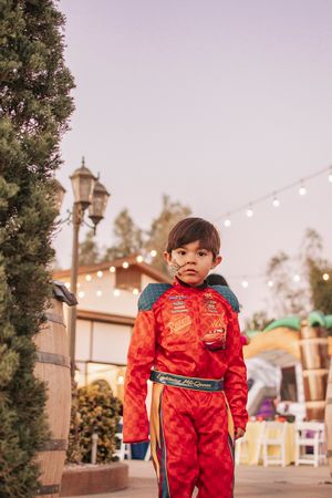 Child wearing bright red racing outfit stands outdoors under string lights and lampposts at dusk.