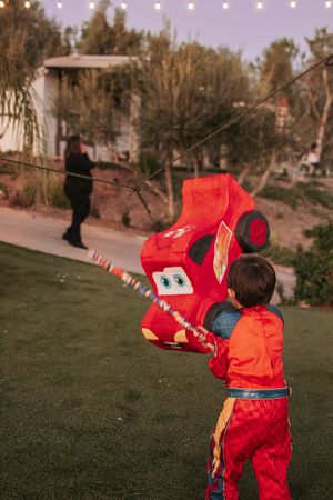 Child in red racing suit playing with a large Cars-themed piñata in a backyard.