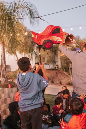 Group of kids watching a red car piñata being hit at an outdoor party with string lights.