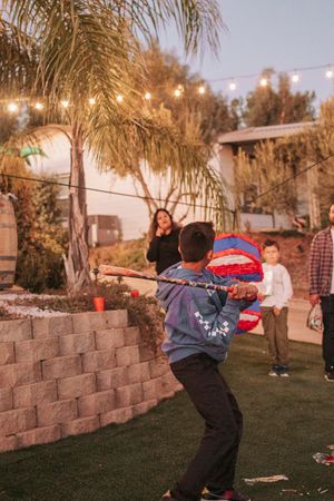 Child swinging at a red piñata in backyard party setting with string lights.