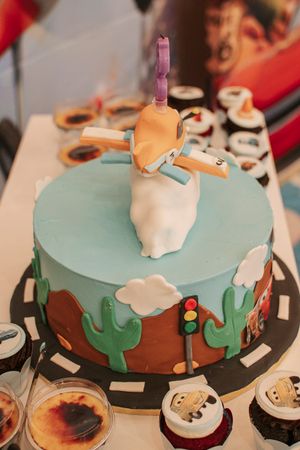 A themed birthday cake decorated with cacti and clouds on a mint green and brown background with cupcakes nearby.
