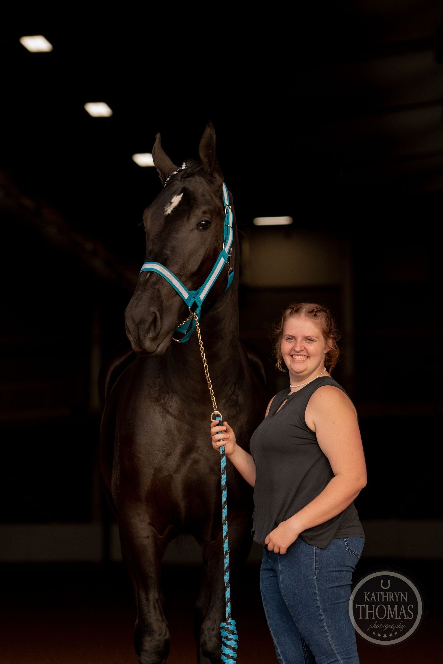 A dark horse wearing a bright turquoise halter stands with its handler in an indoor equestrian setting.