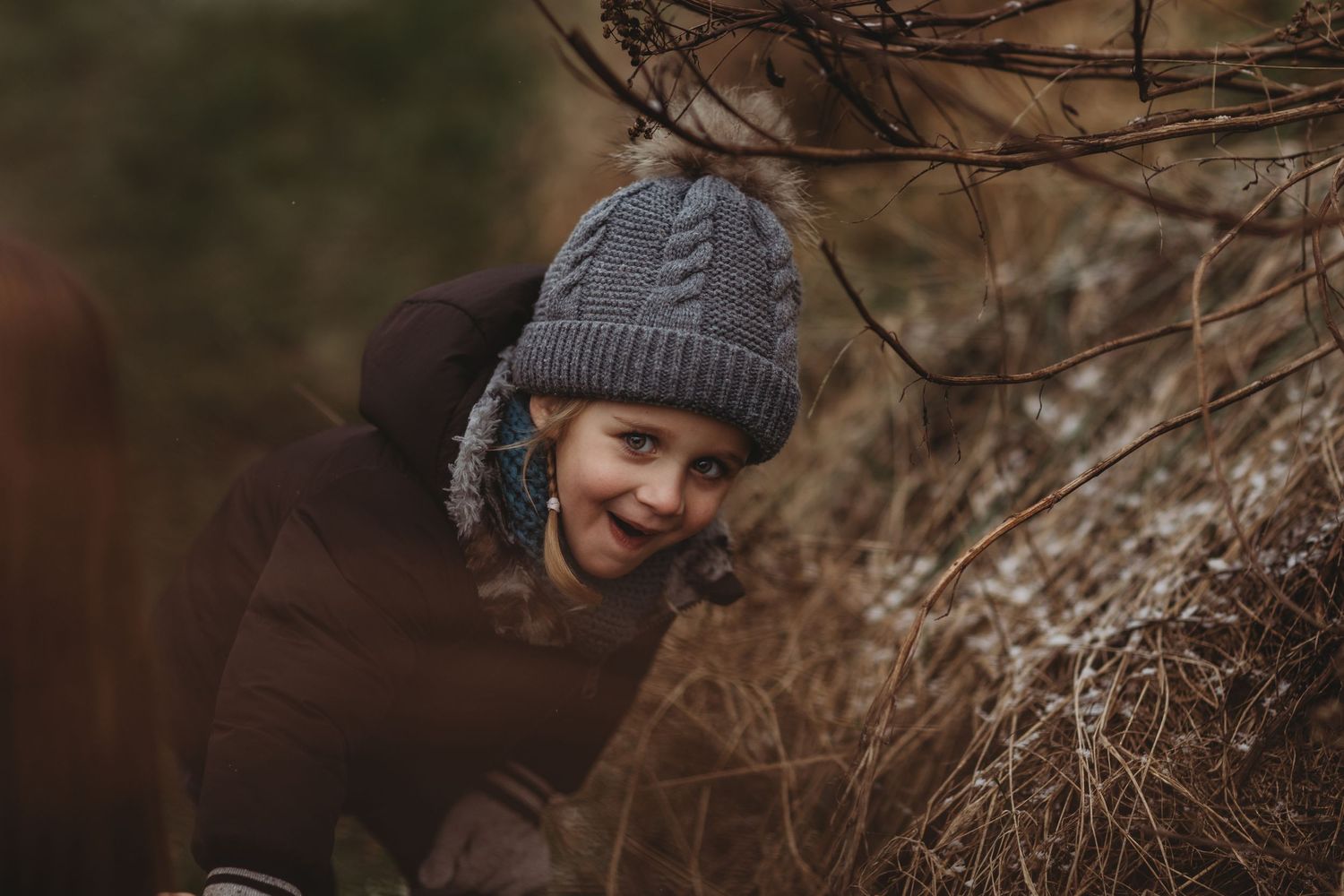 A cozy winter portrait featuring someone wearing a gray knit beanie and brown coat among bare branches.