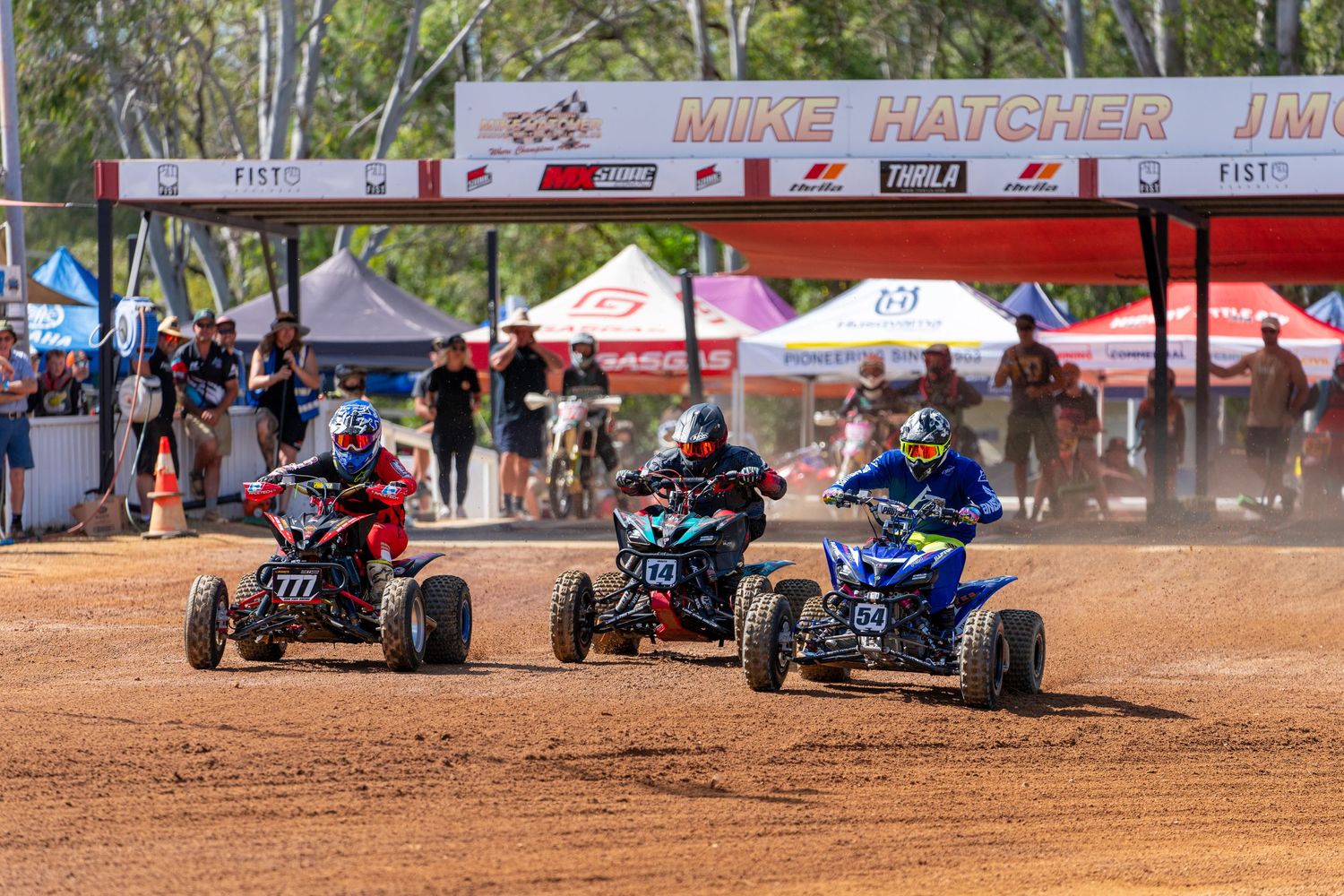 Three ATV riders race neck-and-neck on a dusty dirt track.