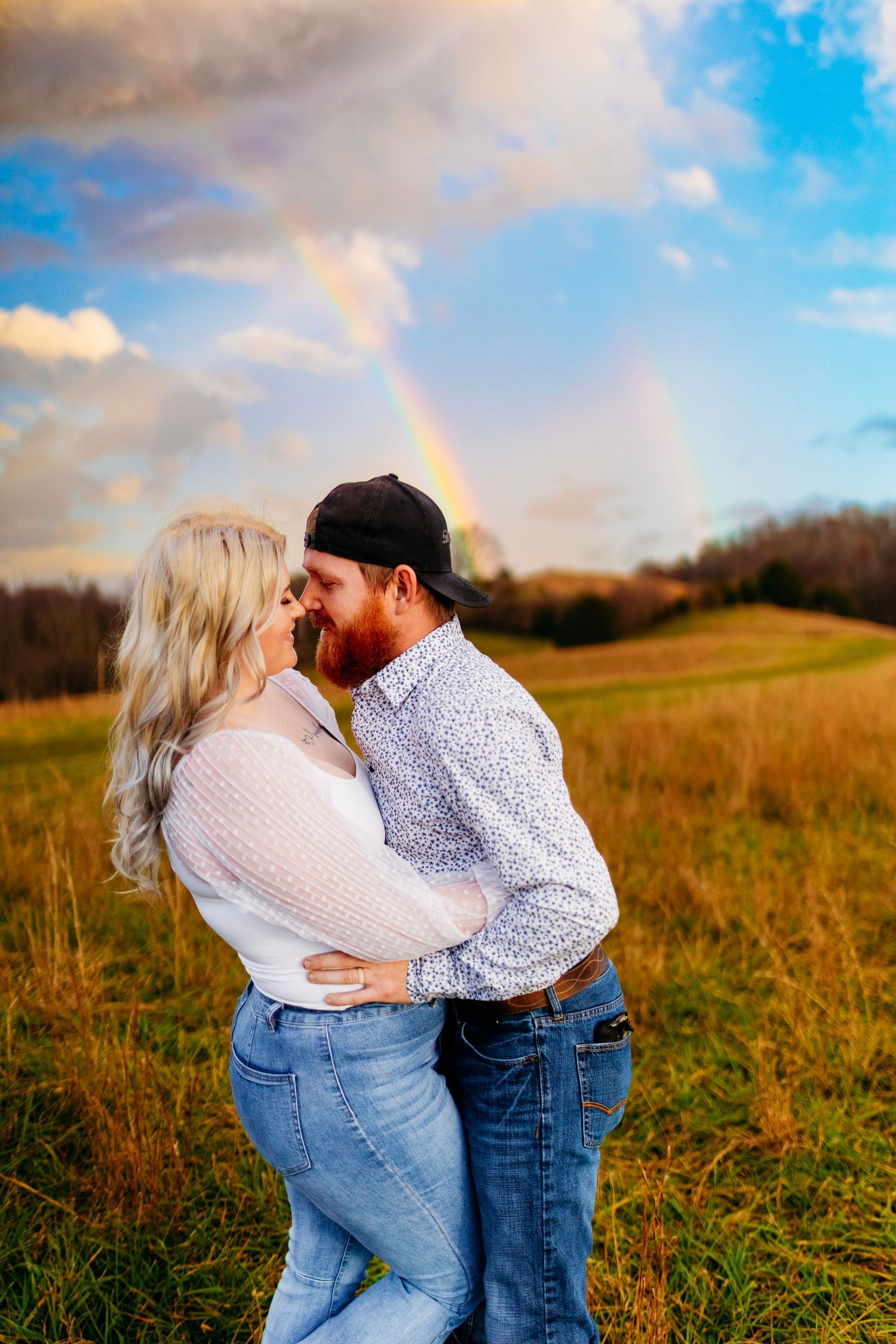 Couple embracing in a field with a rainbow in the sky during golden hour.