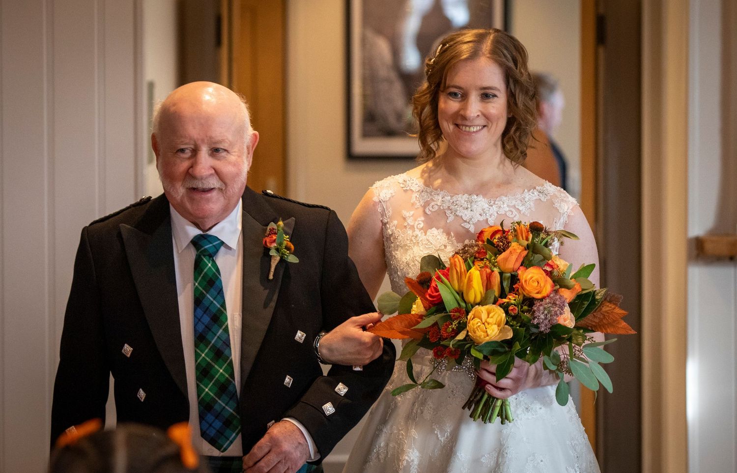 A wedding party member in a green tartan tie walks with a bride holding a vibrant orange and yellow bouquet.