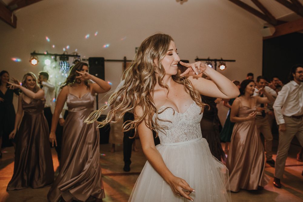 A bride dancing with joy at her reception while bridesmaids in brown dresses celebrate behind her.
