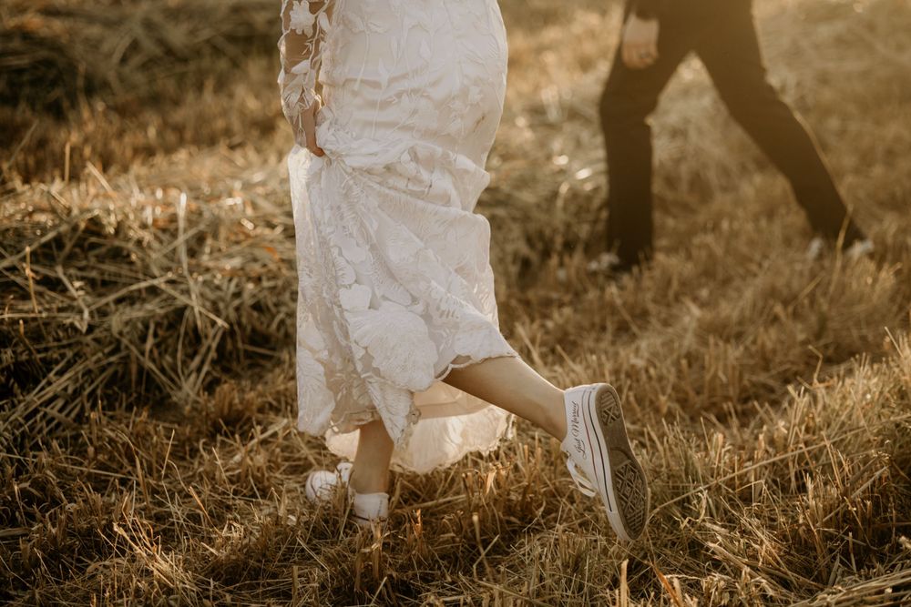 Close-up of white wedding dress and casual sneakers on grassy field during sunset.