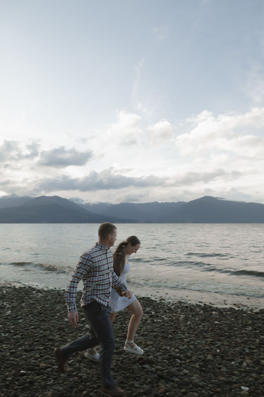 Couple running playfully along a rocky beach with mountains and cloudy skies in the background.